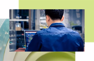 Rear view of a man in a blue work uniform using a laptop in an industrial setting.