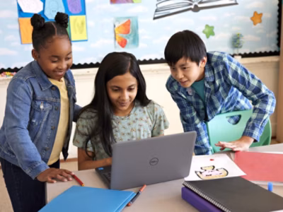 Three children gather around a laptop in a classroom, smiling and engaged. Colorful artwork decorates the wall behind them, and books and drawings are on the table.