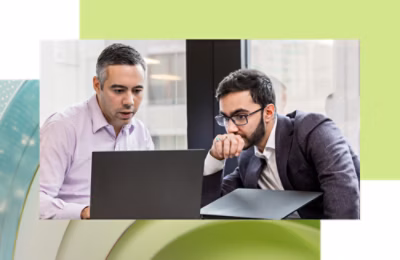 Three people sitting at a table with laptops in a library or office, engaging in a video conference call displayed on a monitor.