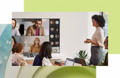 A woman presents to colleagues in a conference room with a large screen showing remote participants in a video call.
