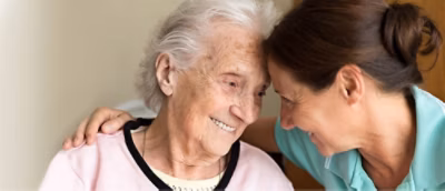 A caregiver smiling affectionately at an elderly woman who is also smiling, in a warm indoor setting.