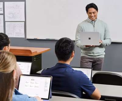 A teacher smiles while holding a laptop, addressing students in a classroom.