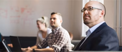 Three people sit attentively in a meeting room. A man in the foreground wears glasses and a suit.
