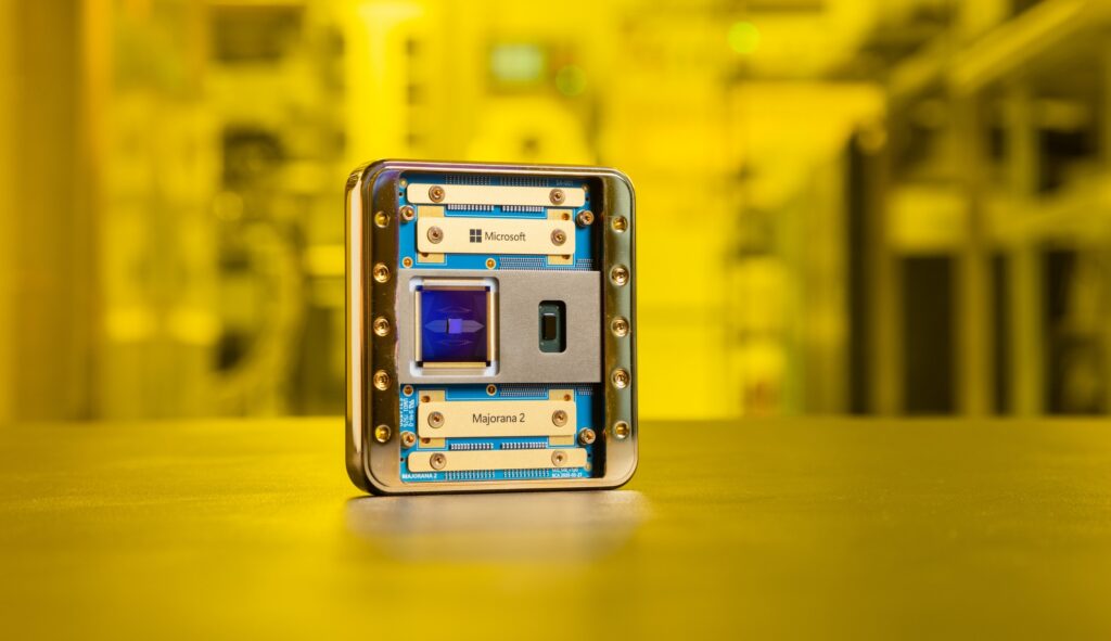 A mostly gold-colored chip with bits of blue showing stands on a table top against a yellow-hued lab background. The names "Microsoft" and "Majorana 2" are visible.